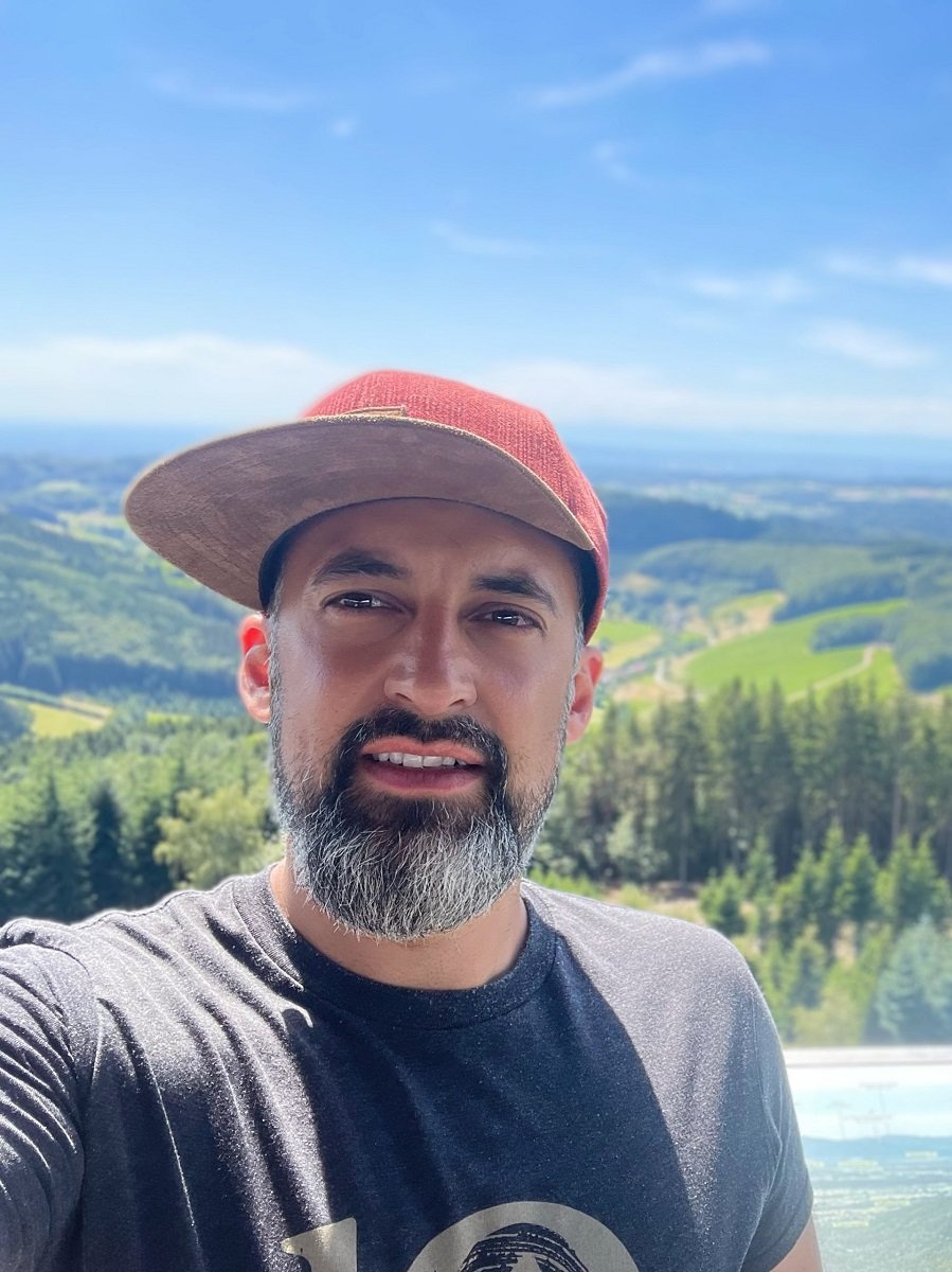 Selfie of Dominik Witzigmann in a baseball cap on a sunny day with a panoramic view of valleys and mountains behind him